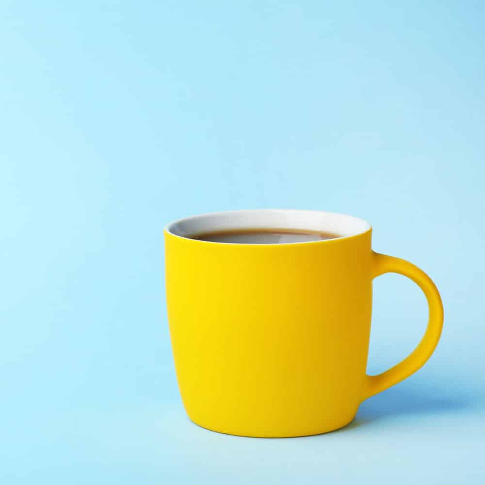 alt=„eine gelbe Henkeltasse mit Kaffee steht vor blauem Hintergrund“