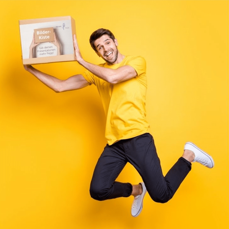alt=„Mann mit gelbem T-Shirt springt in die Luft und hält Bilder-Kiste in beiden Händen“